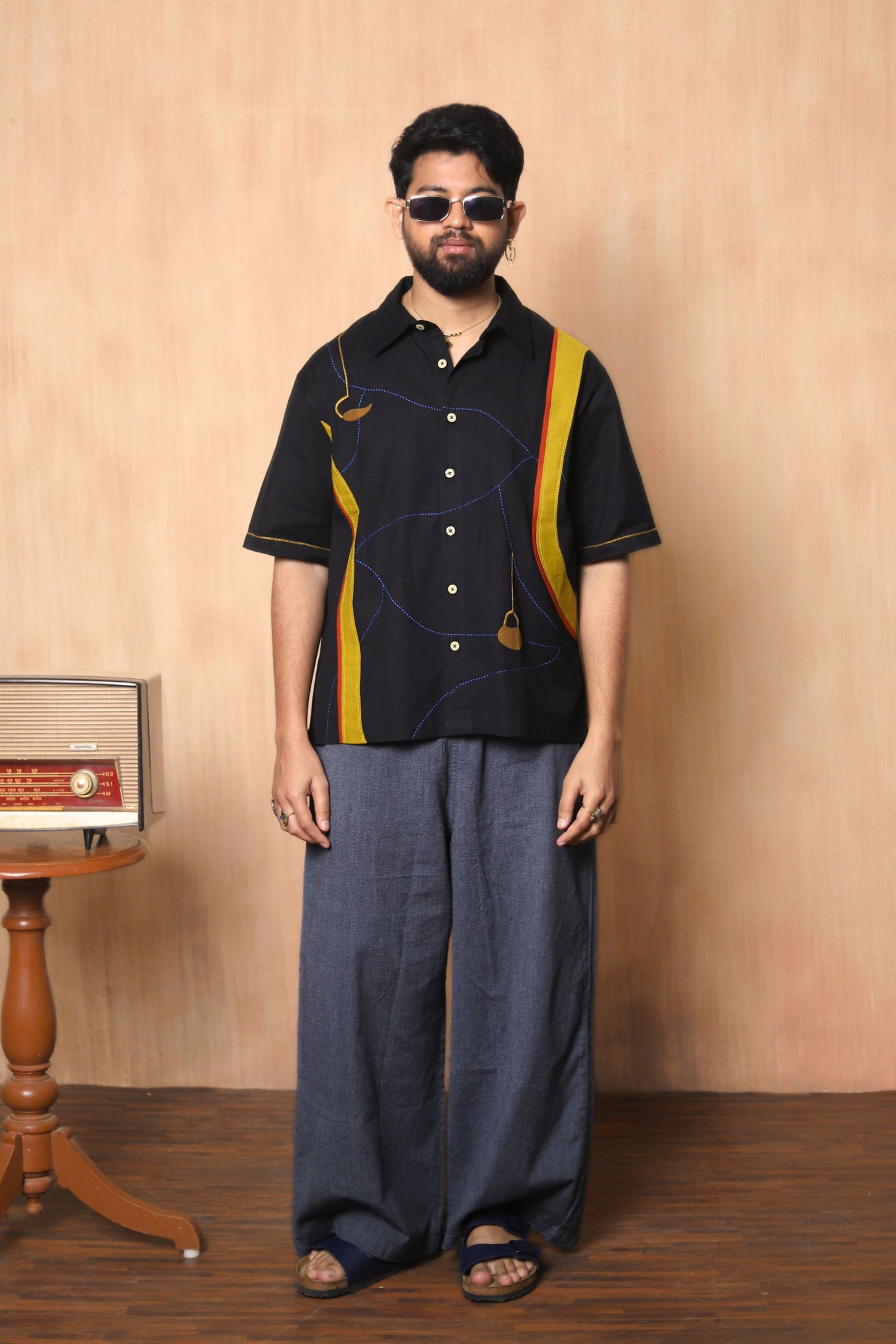 Man wearing a black shirt with yellow accents and blue pants against a beige wall.