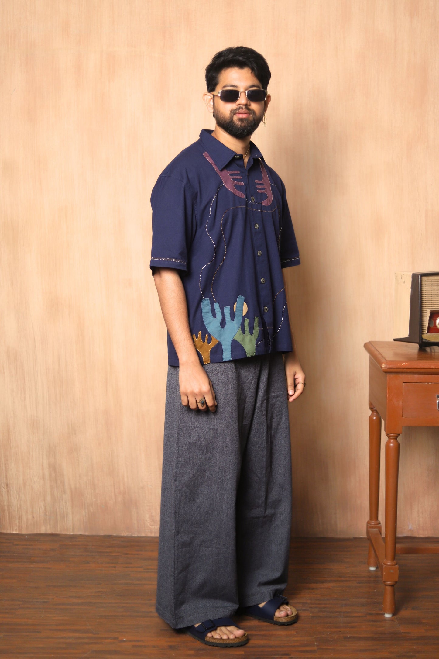 Man wearing a dark blue embroidered shirt and gray wide-leg pants against a beige wall.