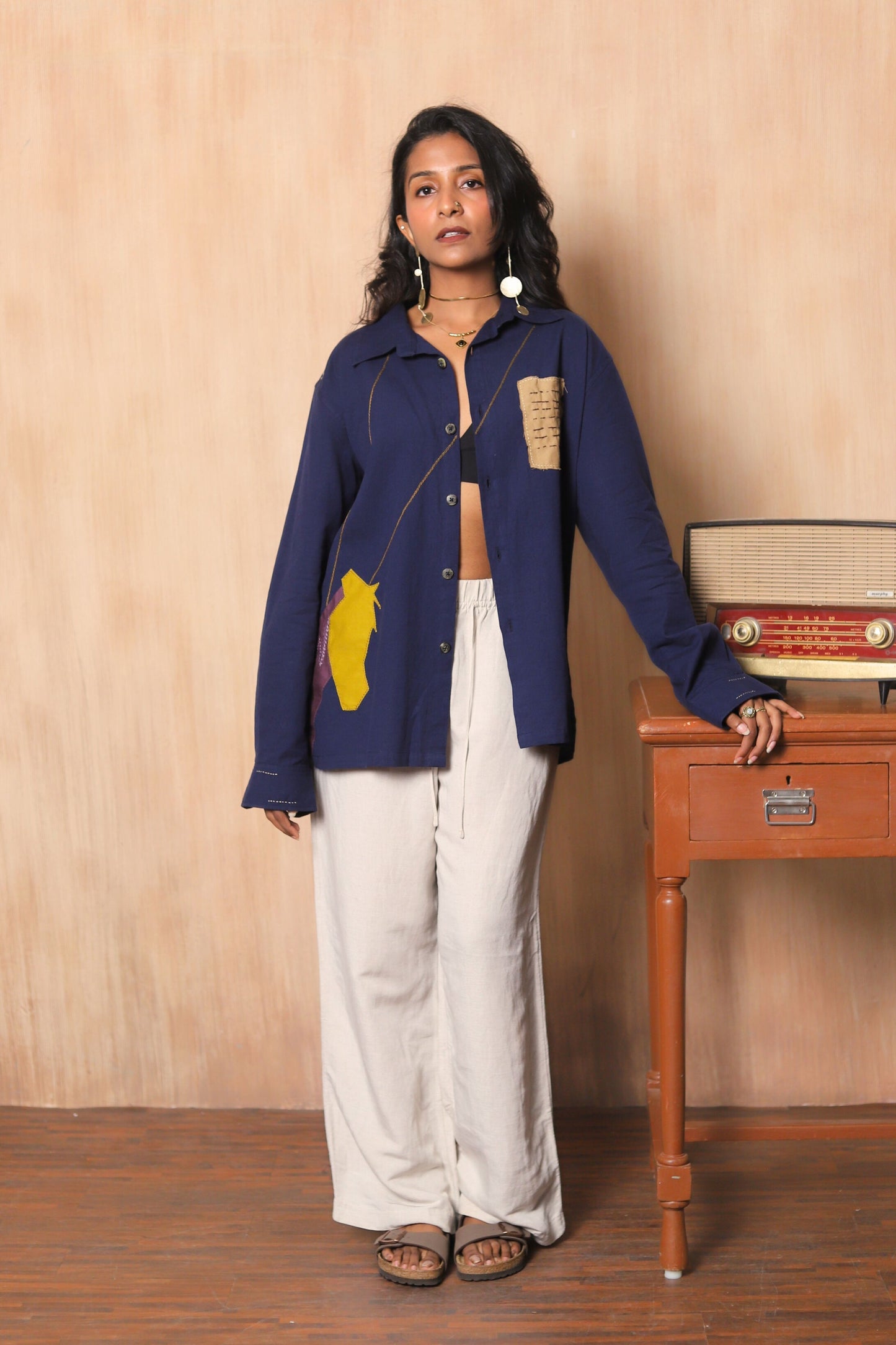 Woman wearing a blue jacket with a yellow patch, standing next to a wooden table with a vintage radio.