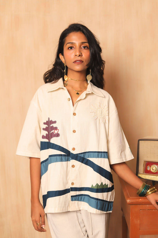 Woman wearing a white shirt with blue stripes and a floral design, standing against a beige wall.
