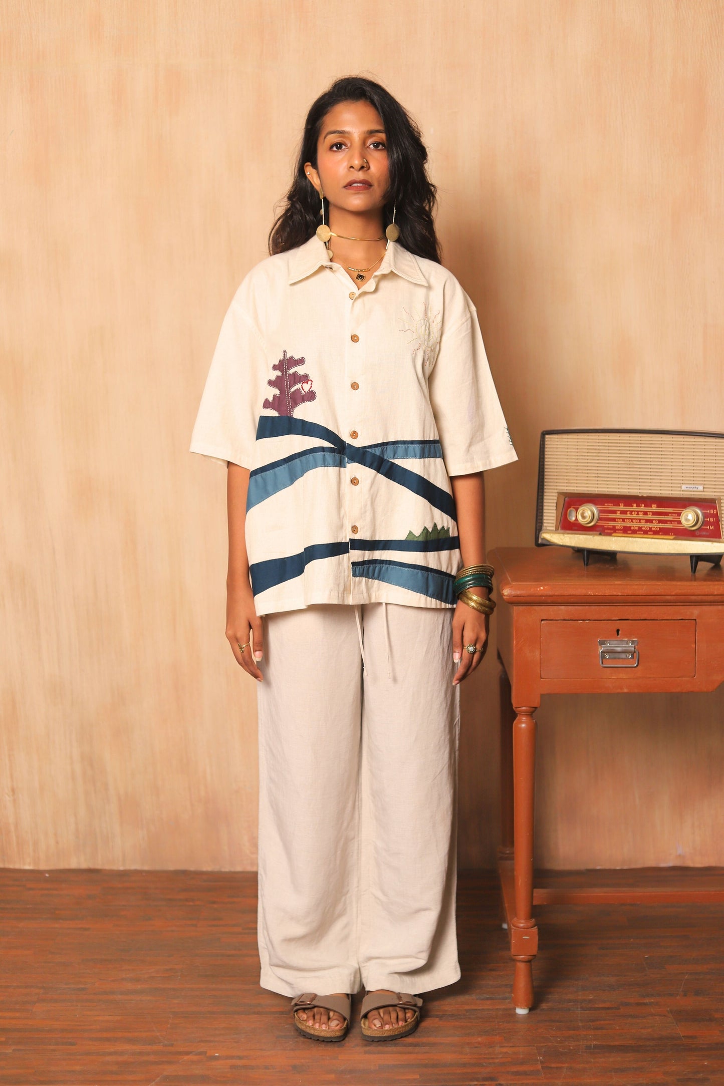 Woman wearing a white shirt with blue stripes and white pants standing next to a wooden table with a radio on it.