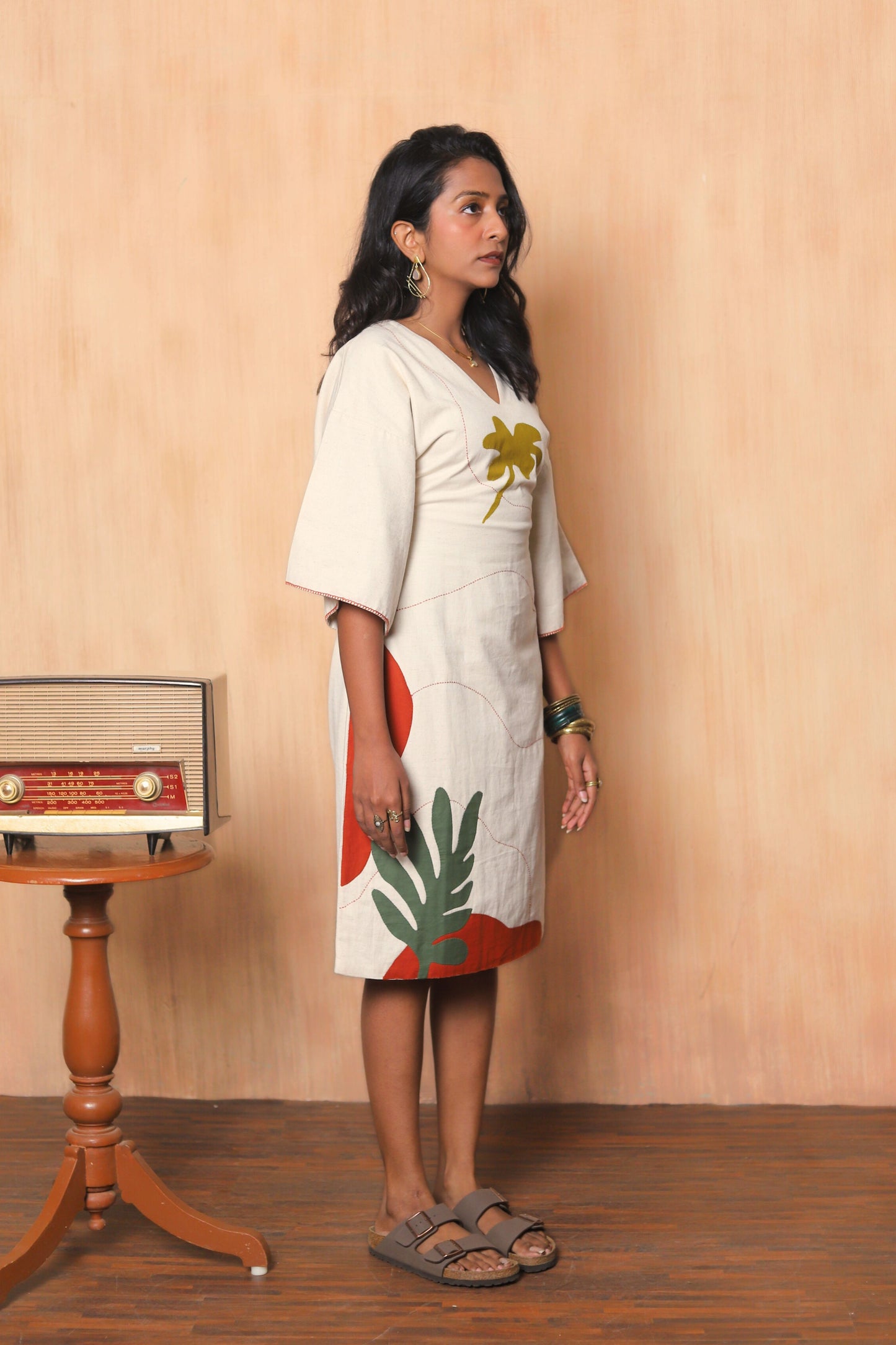Woman wearing a white dress with colorful leaf patterns standing in front of a beige wall.