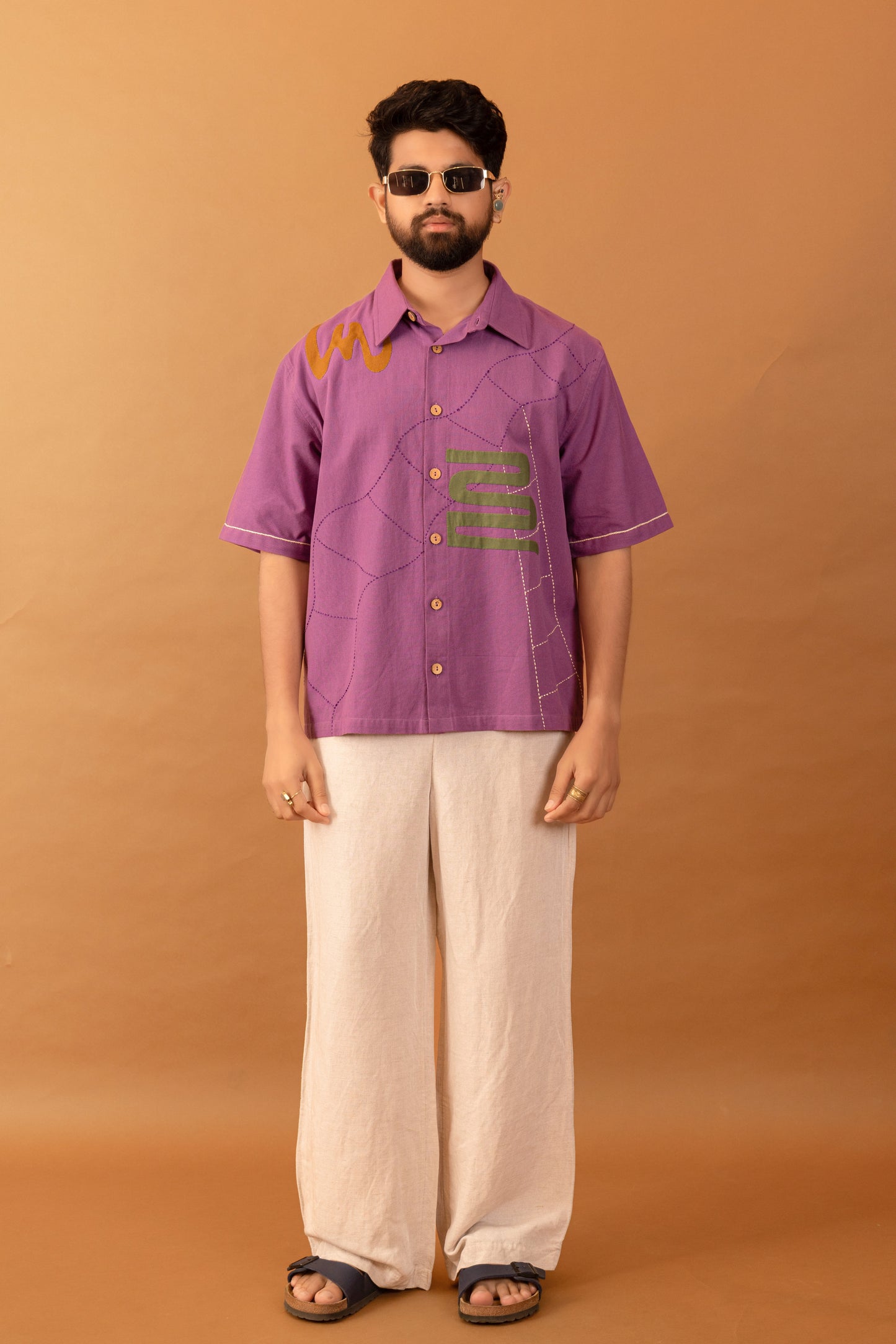 Man wearing a purple shirt with text and white pants against an orange background