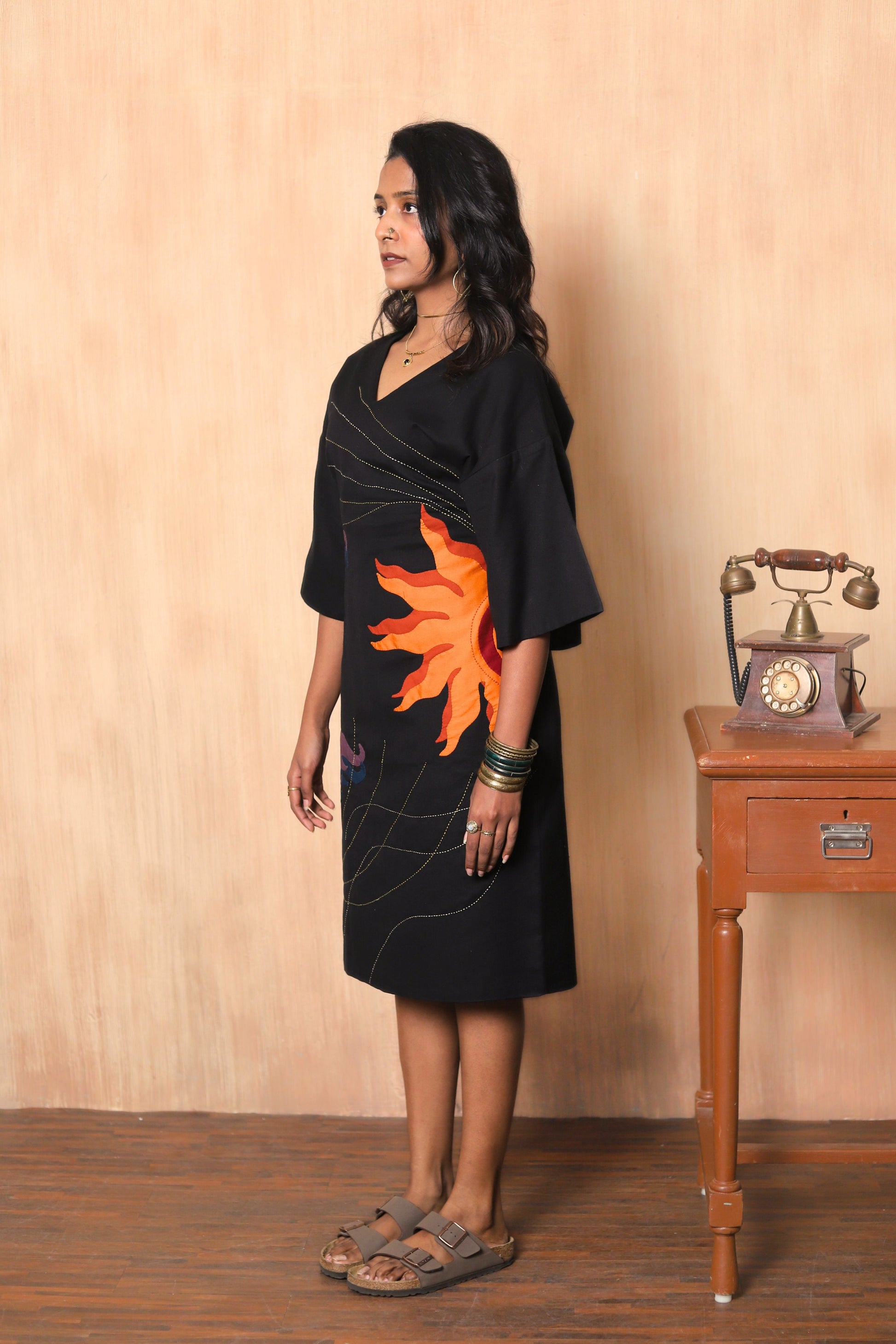 Woman wearing a black dress with a sunflower design in a room with a wooden table and vintage phone.
