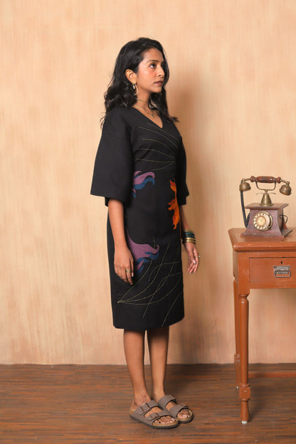 Woman in a black dress with colorful patterns standing next to an old-fashioned telephone on a wooden table.