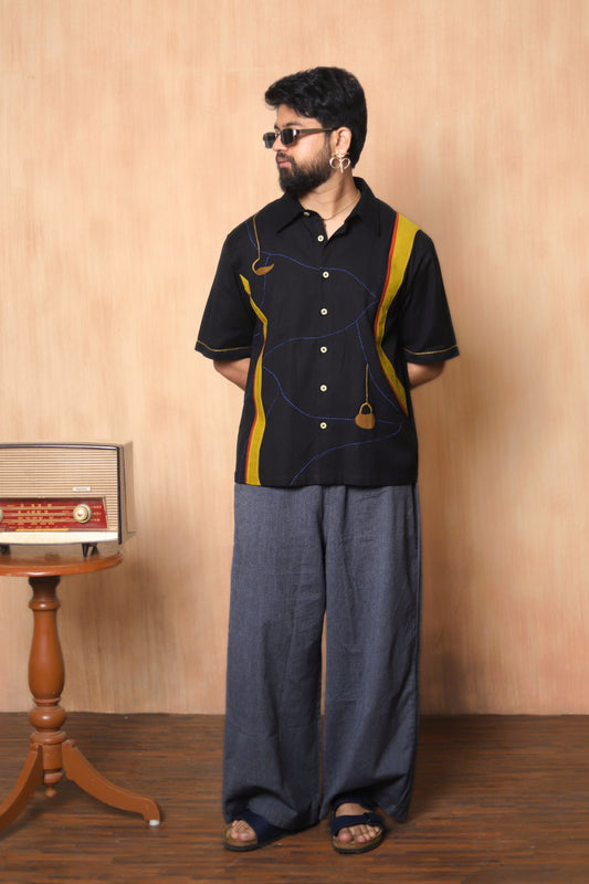 Man wearing a black shirt with yellow accents and blue pants standing in a room with wooden floor and wall.