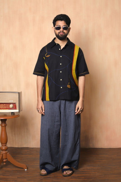 Man wearing a black shirt with yellow accents and blue pants against a beige wall.