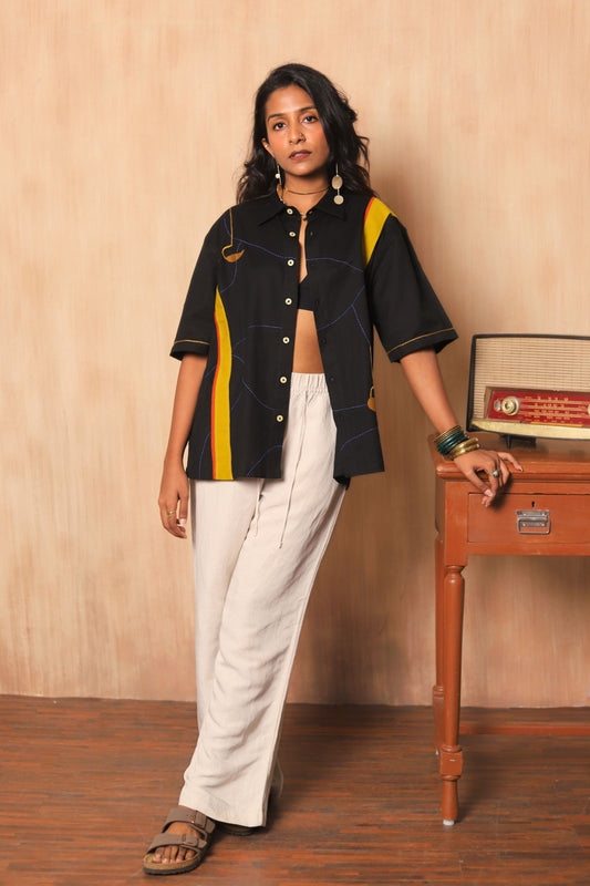 Woman wearing a black jacket with colorful stripes and white pants standing in front of a wooden table with a radio.