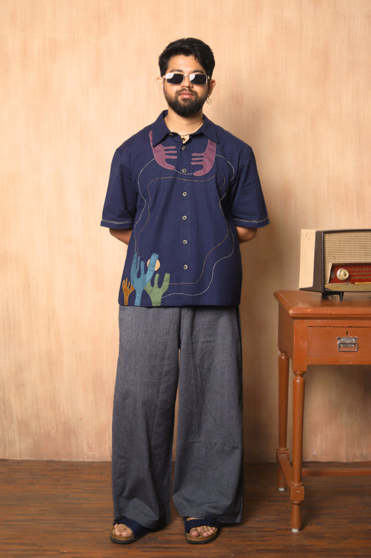 Man wearing a dark blue shirt with colorful designs and gray pants standing against a beige wall.