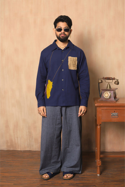 Man wearing a blue shirt with a yellow patch and gray pants standing in front of a wooden wall with a vintage phone.