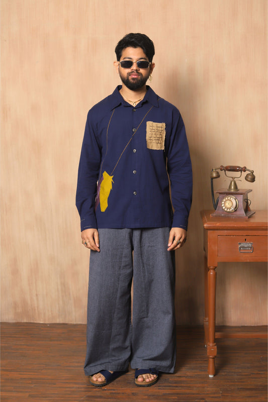 Man wearing a blue shirt with a yellow patch and gray pants standing in front of a wooden wall with a vintage phone.