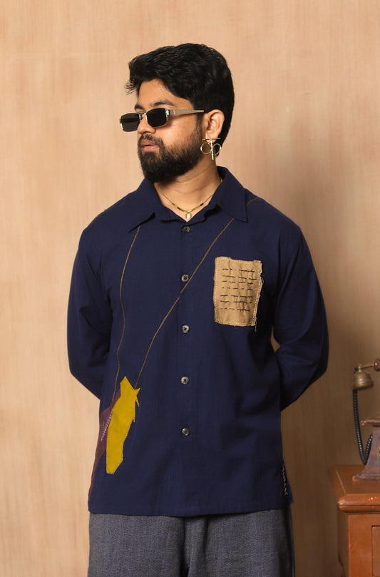 Man wearing a navy blue shirt with a yellow pocket square and gray pants, standing against a wooden paneled wall.
