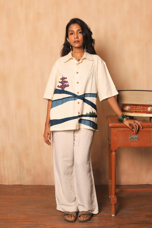 Woman wearing a patterned shirt and white pants standing next to a wooden radio against a beige wall.