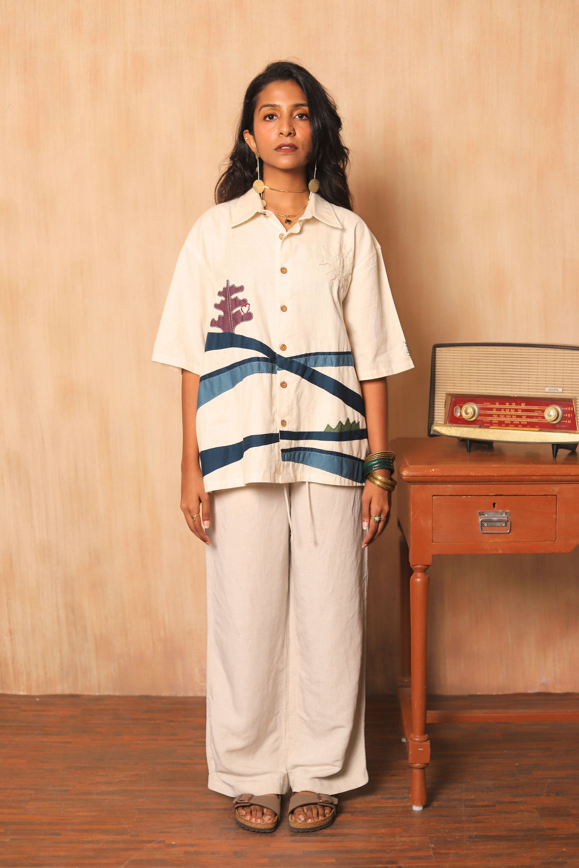 Woman wearing a white shirt with blue stripes and white pants standing next to a wooden table with a radio on it.