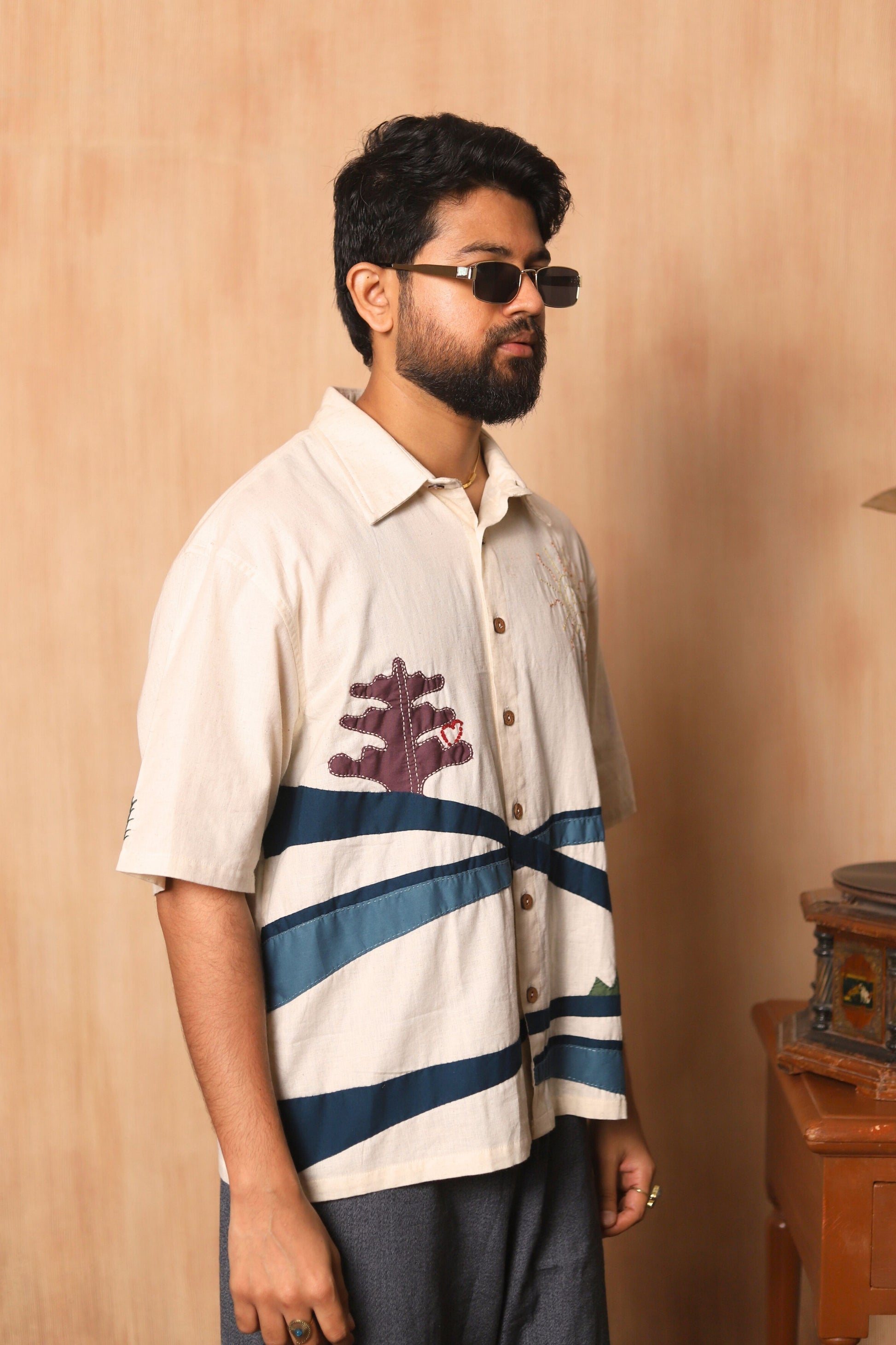 Man wearing a white shirt with blue stripes and a graphic design, standing against a beige wall.
