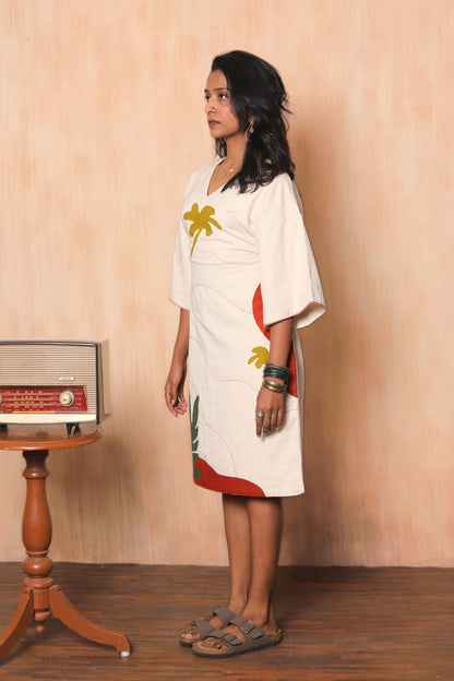 Woman wearing a white dress with colorful designs standing against a wooden wall.