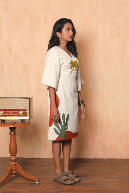 Woman wearing a white dress with colorful leaf patterns standing in front of a beige wall.